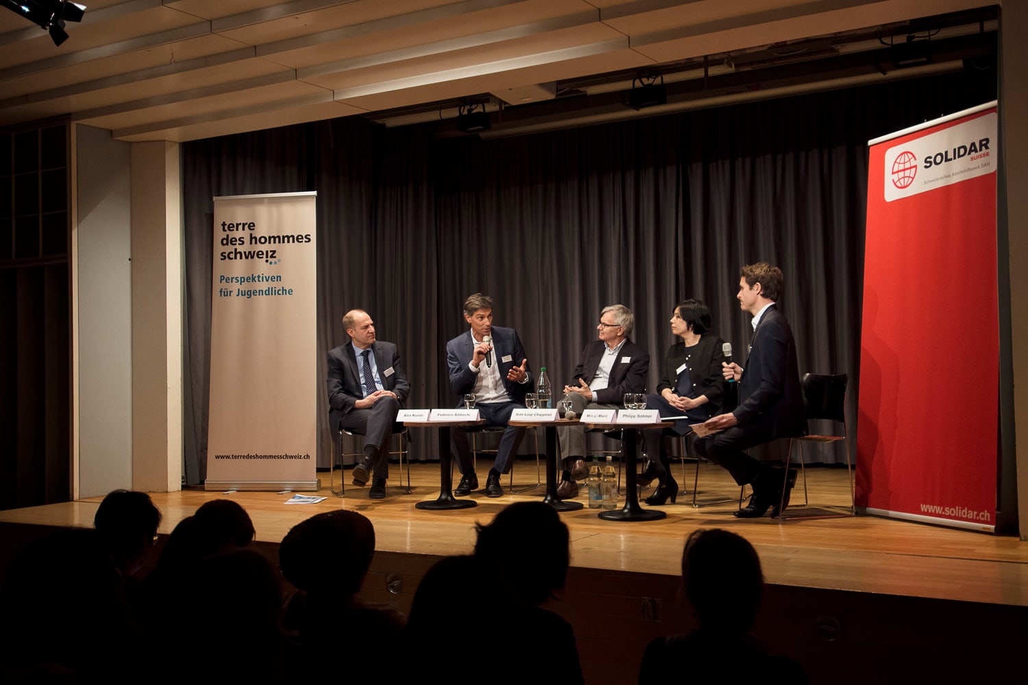 Photo of the five podium participants on stage. Heads of the audience in the foreground.