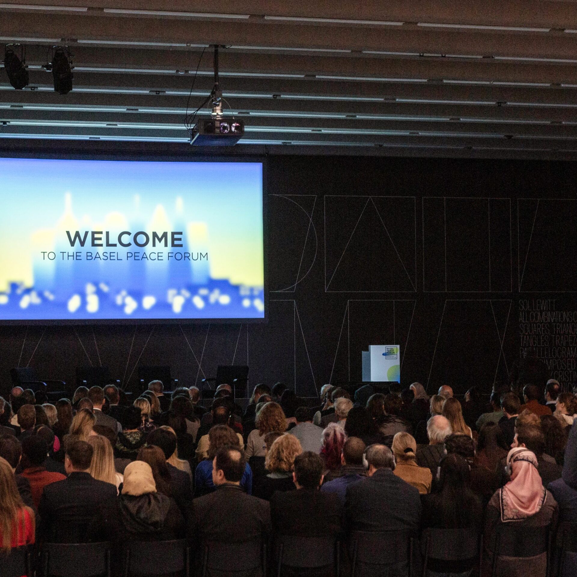 Canvas with beamer picture: Welcome to the Basel Peace Forum. Audience in the darkened hall.